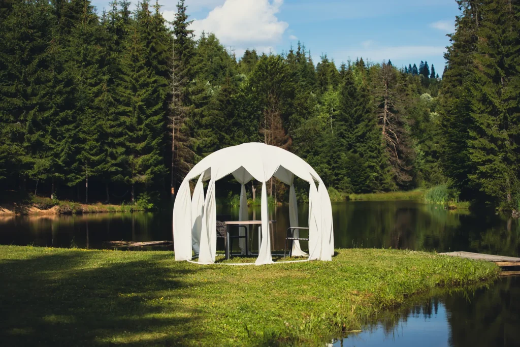 Gazebo blanco con mesa y sillas, situado junto a un lago rodeado de árboles verdes en un día soleado. Ideal para eventos al aire libre y momentos de relajación en la naturaleza.