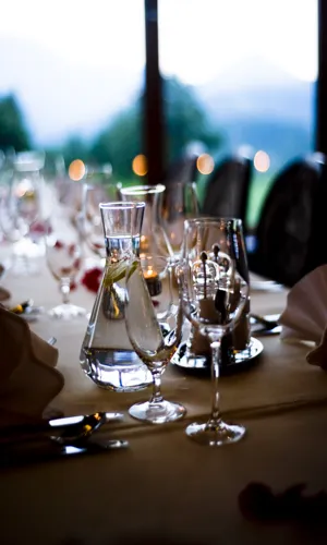 Mesa elegantemente decorada para una cena, con copas de cristal, una jarra de agua y velas, rodeada de un ambiente natural y relajante.