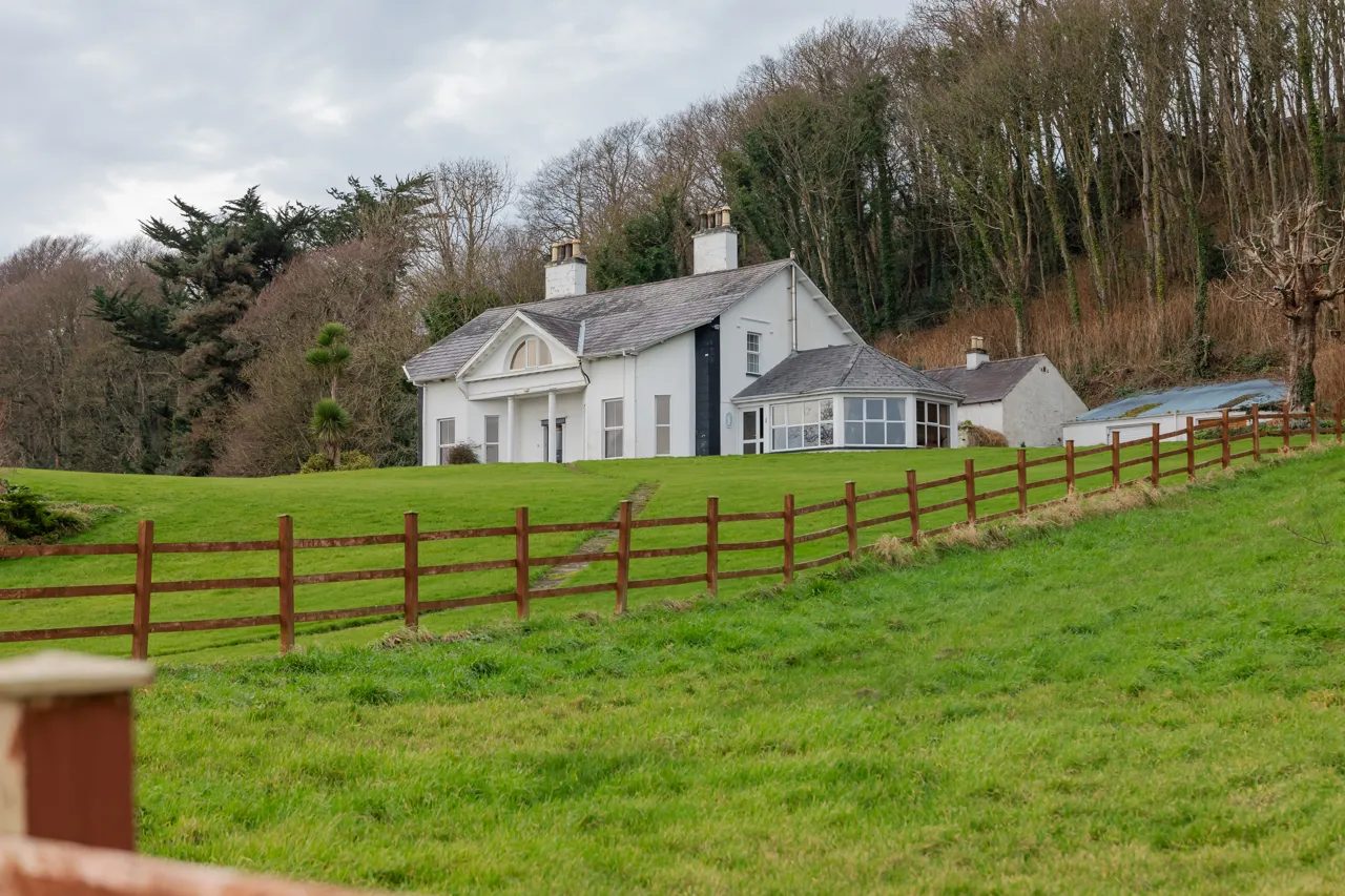 Casa blanca de estilo clásico ubicada en un campo verde, rodeada de un cercado de madera y con árboles al fondo. Ideal para mostrar arquitectura rural y paisajes naturales.