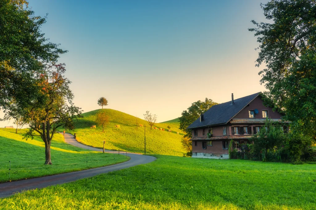 Casa de campo rodeada de colinas verdes y un camino serpenteante al atardecer, con un cielo despejado y árboles en el entorno natural.